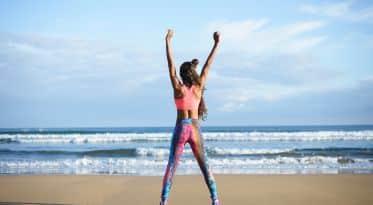 Nainen urheiluvaatteissa katsoo rannalta merelle ja on nostanut kädet ilmaan.